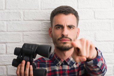 Genç yetişkin adam dürbün kamera ve size parmak ile işaret bakarak tuğla duvarın üzerinden, el işareti, pozitif ve kendinden emin bir jest ön