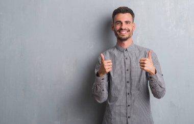 Genç yetişkin adam gri grunge duvar başarı işareti el ile pozitif jest yapıyor üzerinde ayakta başparmak gülümseyen ve mutlu. Neşeli bir ifadeyle, kazanan jest kameraya bakıyor.