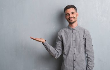 Genç yetişkin adam neşeli sunulması ve kameraya bakarak avucunun ile işaret gülümseyen gri grunge duvarın üzerinden ayakta.