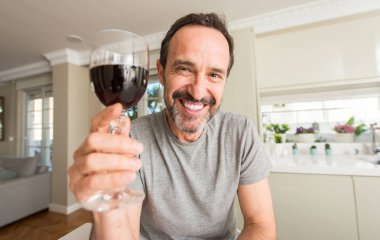 Orta yaş adam ayakta ve diş gösterilen kendine güvenen bir gülümsemeyle gülümseyerek mutlu bir yüz ile bir kadeh şarap içmek