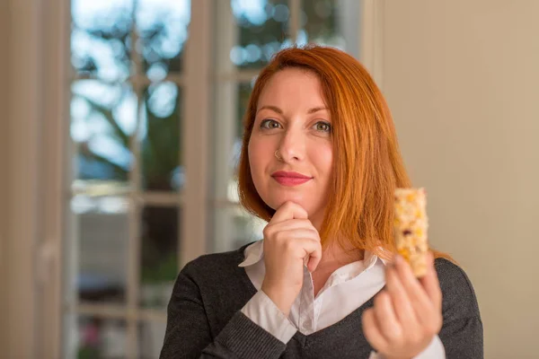 Redhead Woman Eating Cereal Bar Home Serious Face Thinking Question — Stock Photo, Image