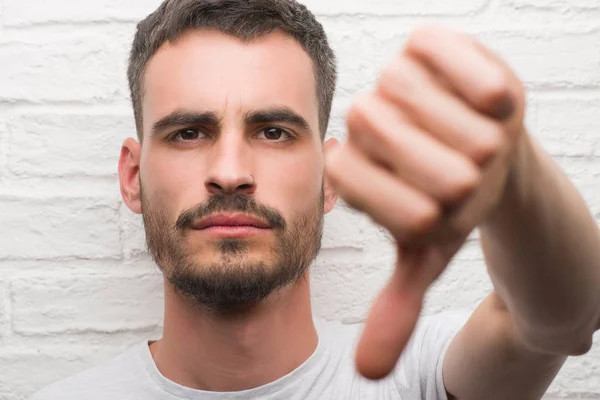 年轻的成年男子站在白色砖墙与愤怒的脸 消极的迹象显示不喜欢拇指向下 拒绝概念 — 图库照片