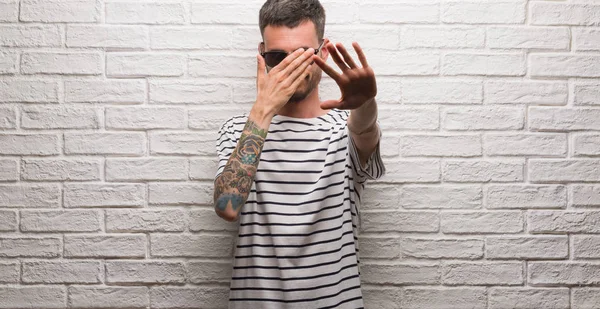 Genç yetişkin adam giyen güneş gözlüğü üzerinde beyaz tuğla ayakta eller gözlerle kaplama duvar ve stop jest ile acı ve korku ifadesi. Utandım ve negatif kavramı.