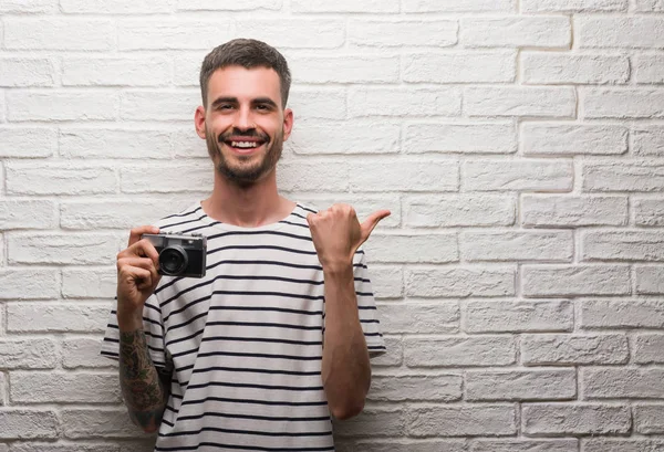 Young man holding vintage camera standing over white brick wall pointing and showing with thumb up to the side with happy face smiling