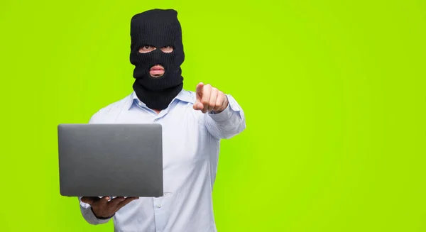 Business hacker man holding a computer laptop pointing with finger to the camera and to you, hand sign, positive and confident gesture from the front