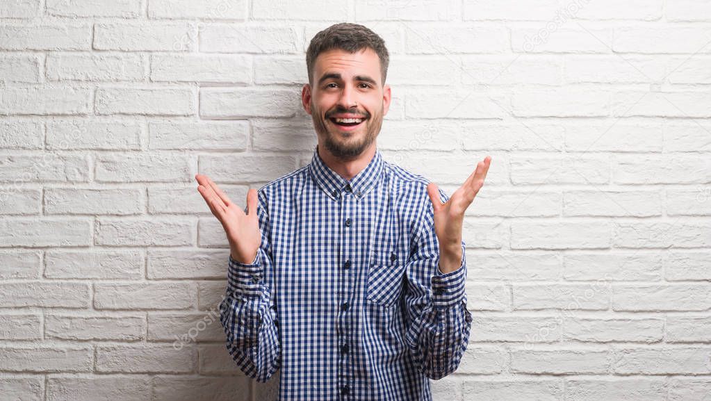 Young adult man standing over white brick wall very happy and excited, winner expression celebrating victory screaming with big smile and raised hands