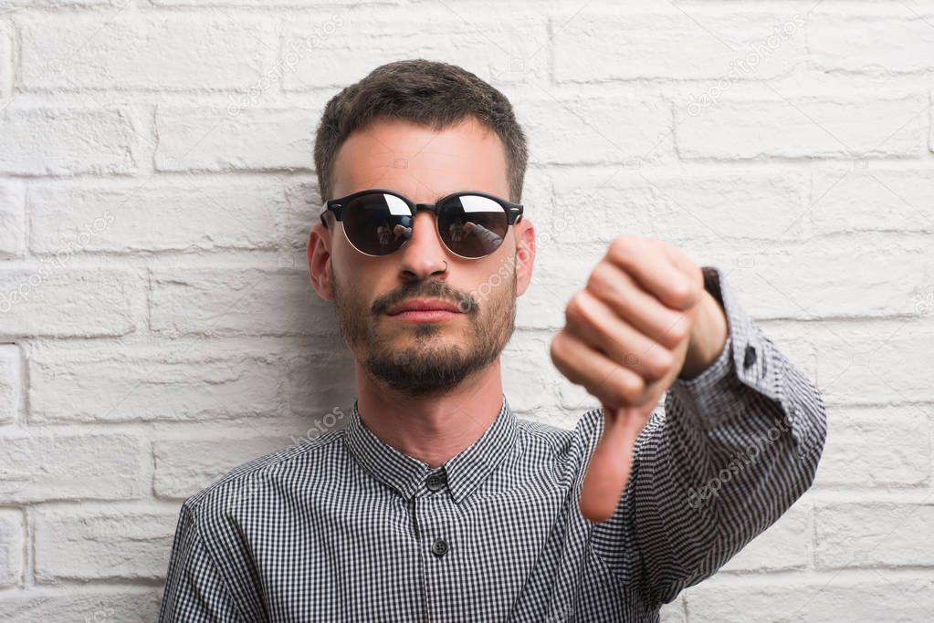 Young adult man wearing sunglasses standing over white brick wall with angry face, negative sign showing dislike with thumbs down, rejection concept