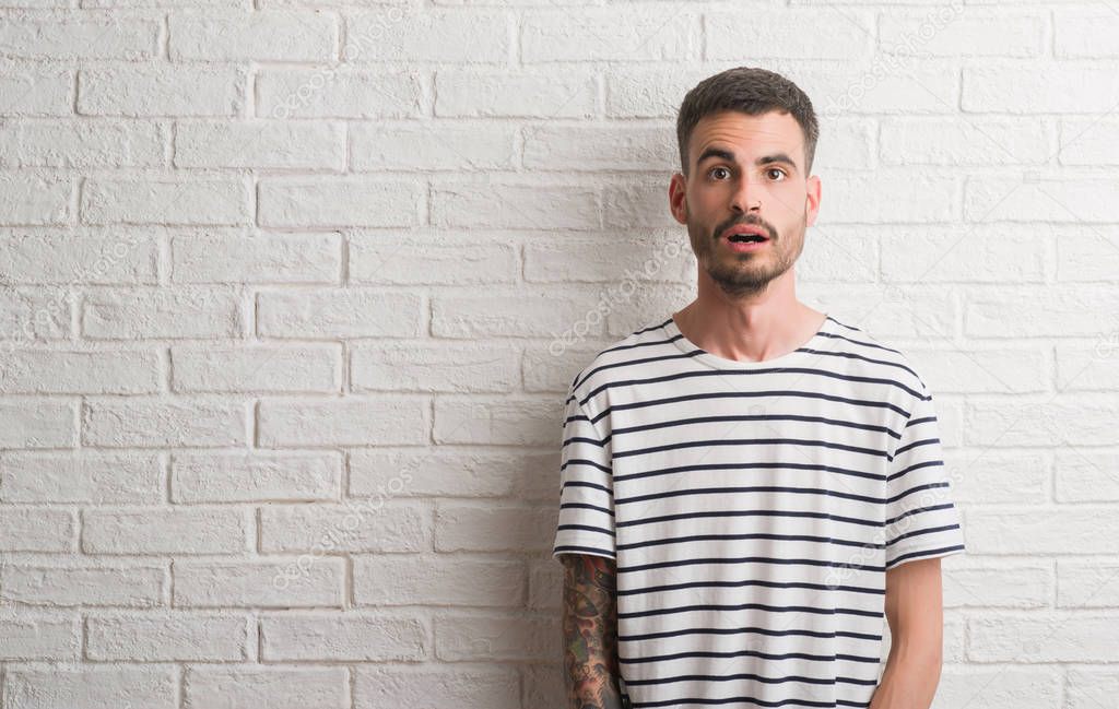 Young adult man standing over white brick wall scared in shock with a surprise face, afraid and excited with fear expression