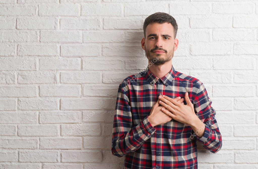 Young adult man standing over white brick wall smiling with hands on chest with closed eyes and grateful gesture on face. Health concept.