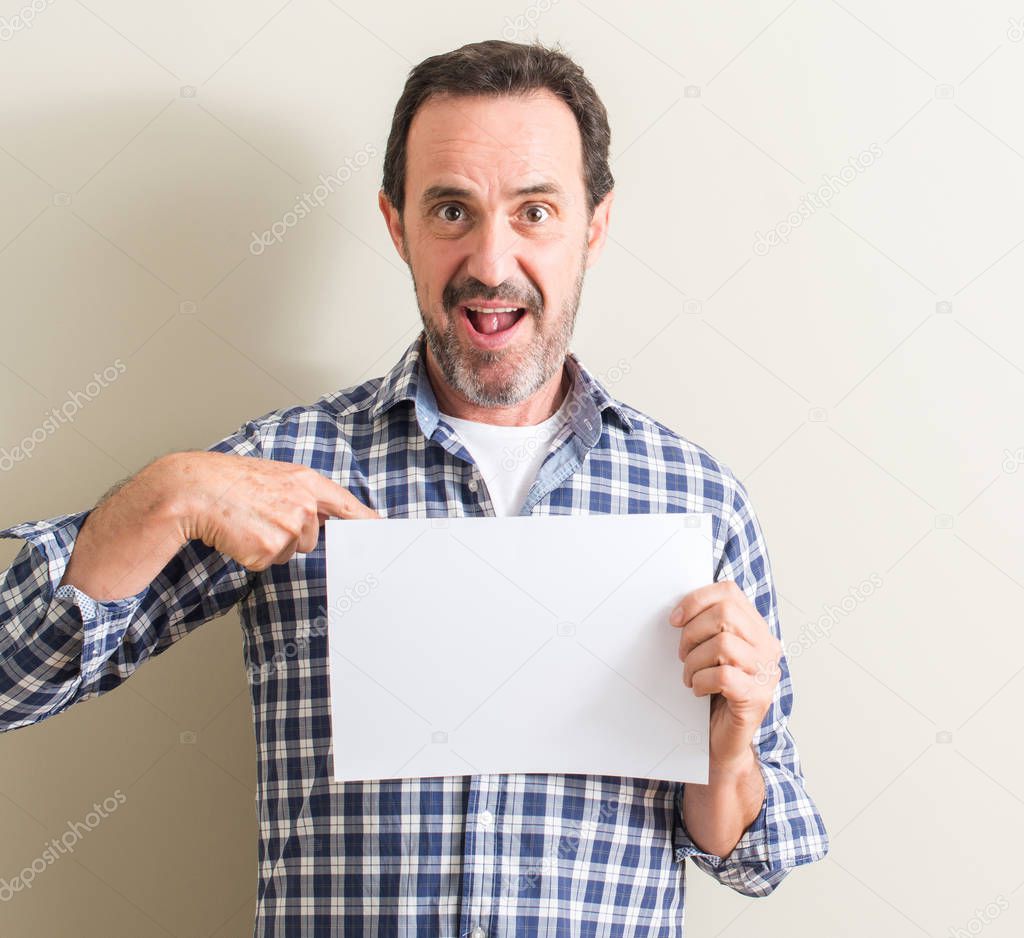 Senior man holding blank paper sheet with surprise face pointing finger to himself