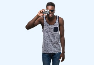 Vintage fotoğraf makinesi ile ayakta ve diş gösterilen kendine güvenen bir gülümsemeyle gülümseyerek mutlu bir yüz tutan genç Afro-Amerikan adam