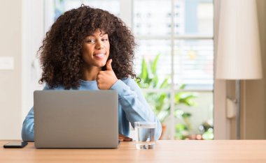 Dizüstü bilgisayar kullanan Afrikalı-Amerikalı kadın evde kocaman bir gülümseme ile ok işareti yaparken mutlu, parmaklarıyla, mükemmel işaret parmak