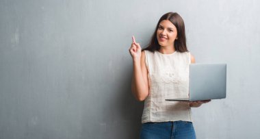 Grunge genç esmer kadın bilgisayar dizüstü sürpriz bir fikir kullanarak duvar gri veya gülen yüz, bir numara ile işaret parmağı soru