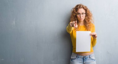 Gri grunge duvar boş kağıt sayfası kamera ve size parmak ile işaret tutan genç Kızıl saçlı kadın, el işareti, pozitif ve kendinden emin bir jest ön