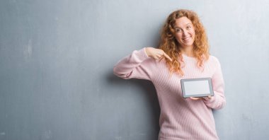 Kendisi için sürpriz yüz işaret parmağı ile tablet kullanarak gri grunge duvar genç Kızıl saçlı kadın