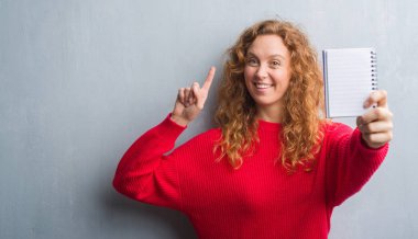 Gri grunge genç Kızıl saçlı kadın boş defter sürpriz bir fikir ile gösterilen duvar veya gülen yüz, bir numara ile işaret parmağı soru