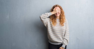 Genç Kızıl saçlı kadın gülümsüyor ve gözleri için sürpriz kapsayan yüz el ile gülüyor gri grunge duvarın üzerinden. Kör kavramı.