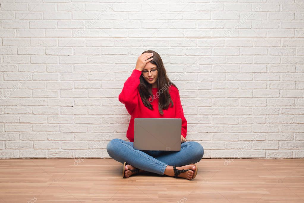 Young brunette woman sitting on the floor over white brick wall using laptop stressed with hand on head, shocked with shame and surprise face, angry and frustrated. Fear and upset for mistake.