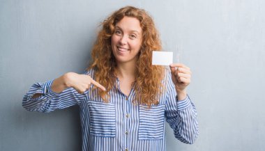 Genç Kızıl saçlı kadın boş tutan gri grunge duvarın üzerinden ziyaret edin kartı sürpriz yüz işaret parmağı kendisine