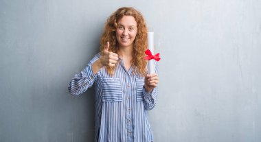 Genç redhead iş kadını ok işareti, başparmak ile parmak, mükemmel işareti yapıyor kocaman bir gülümseme ile diploma mutlu tutarak gri grunge duvarın üzerinden