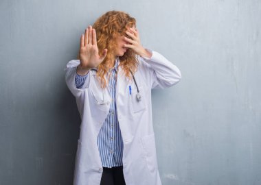 Genç redhead doktor kadın gri grunge duvar eller gözlerle kapsayan bir palto giyiyor ve stop jest ile acı ve korku ifadesi. Utandım ve negatif kavramı.