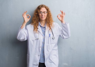 Genç redhead doktor kadın bir palto giyiyor gri grunge duvarın üzerinden rahatla ve gülümseyen gözlerle kapalı parmak jestle meditasyon yapıyor. Yoga kavramı.
