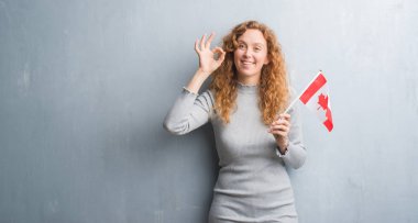 Kanada bayrağı tutarak gri grunge duvarın üzerinden genç Kızıl saçlı kadının parmak, mükemmel sembolü ile ok işareti yapması