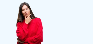 Genç güzel İspanyol emin çapraz kollar ve çeneme kaldırdı el ile gülümseme ile kameraya bakarak kırmızı kazak giyiyor. Pozitif düşünce.