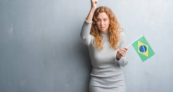 灰色垃圾墙上的年轻红头发的女人拿着巴西国旗恼怒和沮丧的呼喊与愤怒 疯狂和咆哮与举手 愤怒的概念 — 图库照片