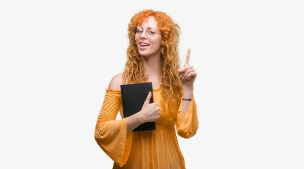 年轻的红发学生妇女拿着一本书惊讶于一个想法或问题指向手指与快乐的脸 — 图库照片