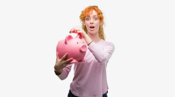 Young Redhead Woman Holding Piggy Bank Scared Shock Surprise Face — Stock Photo, Image