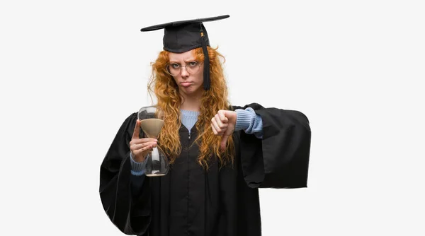 年轻红发学生女子穿着毕业制服持有沙漏与愤怒的脸 否定的标志显示不喜欢拇指向下 拒绝概念 — 图库照片