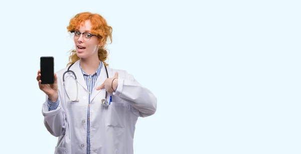 Young redhead doctor woman showing smartphone with surprise face pointing finger to himself