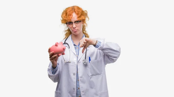 年轻红头发的医生女人抱着小猪银行愤怒的脸 消极的迹象显示不喜欢拇指向下 拒绝概念 — 图库照片
