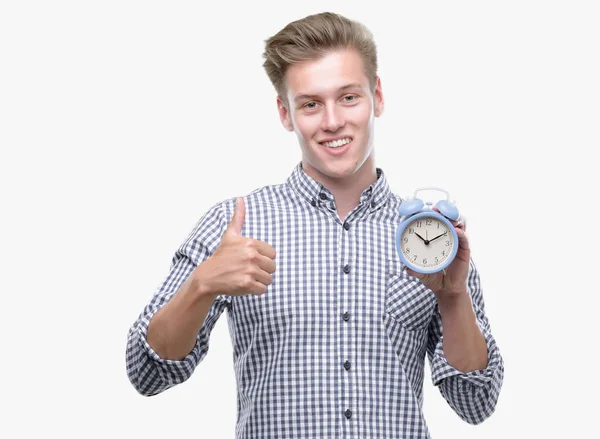 笑顔の サイン 優秀な兆候を親指をやって幸せな目覚まし時計を保持している若いハンサムな金髪男 — ストック写真