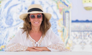 Ayakta ve diş gösterilen kendine güvenen bir gülümsemeyle gülümseyerek mutlu bir yüz ile renkli arka plan ile masada oturan orta yaş esmer kadın