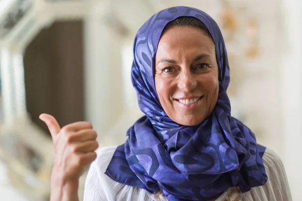 Middle age brunette arabian woman wearing colorful hijab pointing and showing with thumb up to the side with happy face smiling
