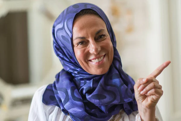 Middle age brunette arabian woman wearing colorful hijab very happy pointing with hand and finger to the side
