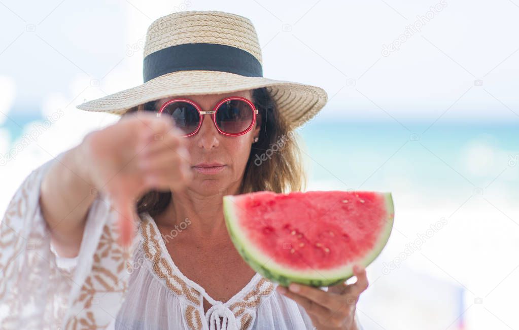 Middle age brunette woman by the sea eating watermelon with angry face, negative sign showing dislike with thumbs down, rejection concept