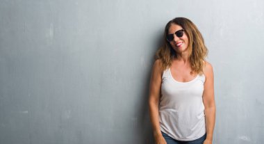 Orta yaş İspanyol kadın gri duvar uzak tarafına yüzünde, doğal ifade gülümseme ile seyir güneş gözlüğü takıyor. Kendine güvenen gülüyor.