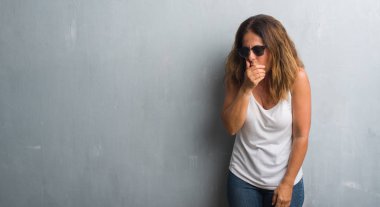 Orta yaş İspanyol kadın gri duvar hasta ve soğuk veya bronşit için semptom olarak öksürük duygu güneş gözlüğü takıyor. Sağlık kavramı.