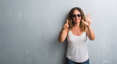 Orta yaş İspanyol kadın gösterilen ve parmakları ile işaret güneş gözlüğü takıyor gri duvar yüzünden kendinden emin ve mutlu gülümseyerek süre 6 numara.