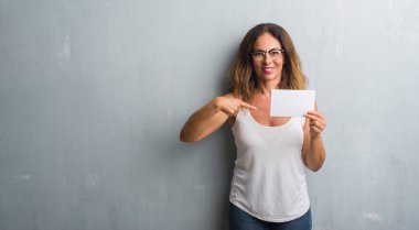 Orta yaş İspanyol kadın kendisine sürpriz yüz işaret parmağı boş kartı tutan gri grunge duvarın üzerinden ayakta