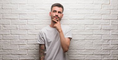 Genç yetişkin adam beyaz tuğla duvar ciddi bir yüz düşünme hakkında soru, çok karışık bir fikir üzerinde ayakta