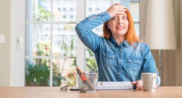 Redhead woman studying at home stressed with hand on head, shocked with shame and surprise face, angry and frustrated. Fear and upset for mistake.