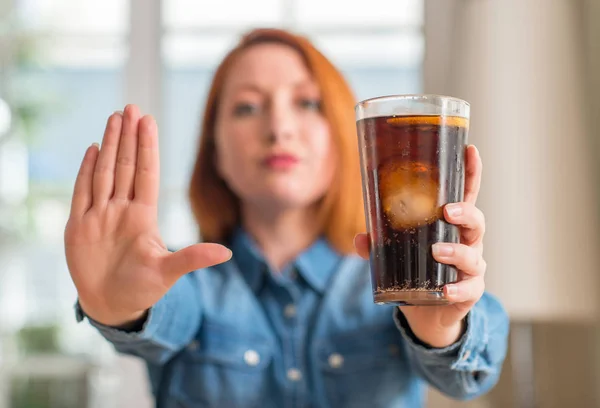 Redhead woman holding soda refreshment with open hand doing stop sign with serious and confident expression, defense gesture