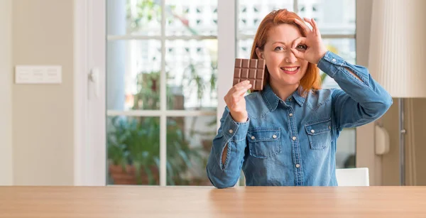 Redhead woman holding chocolate bar at home with happy face smiling doing ok sign with hand on eye looking through fingers