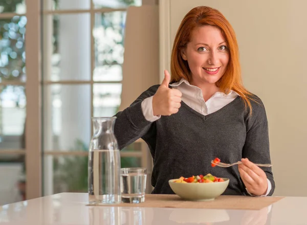 红头发的女人吃水果碗 猕猴桃和草莓在家里开心的大笑做 拇指与手指 优秀的标志 — 图库照片