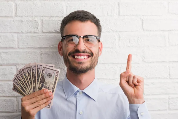 Young hipster business man holding dollars surprised with an idea or question pointing finger with happy face, number one