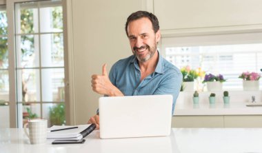 Dizüstü bilgisayar kullanan orta yaş adam evde kocaman bir gülümseme ile ok işareti yaparken mutlu, parmaklarıyla, mükemmel işaret parmak